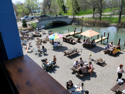 A lively outdoor dining area by a waterway, with people at tables, umbrellas, and a bridge in the background.