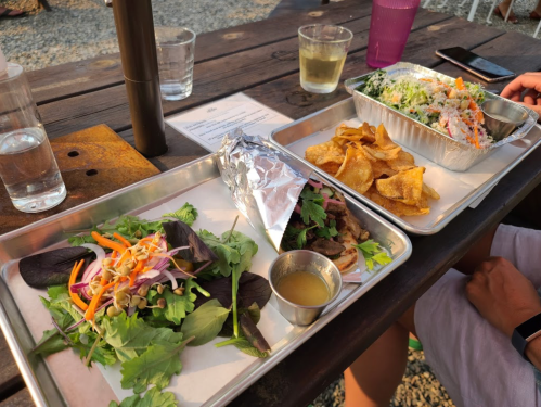 A table with two trays of food: one with a salad and a wrapped item, the other with chips and a seafood dish.