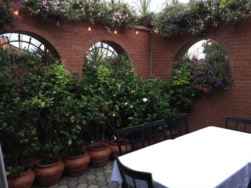 A cozy outdoor dining area with a white table, surrounded by lush greenery and flowering plants in terracotta pots.