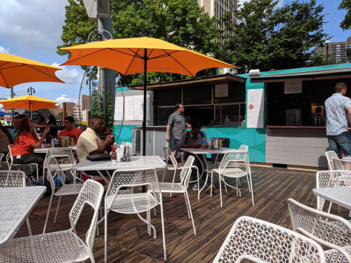 Outdoor café scene with yellow umbrellas, tables, and people dining. A food counter is visible in the background.