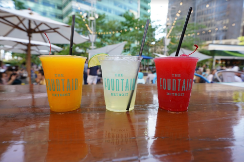 Three colorful drinks in clear cups with straws, sitting on a wooden table at a lively outdoor venue.