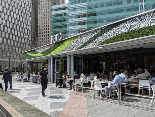 A modern restaurant with outdoor seating, surrounded by tall buildings and greenery, bustling with diners.