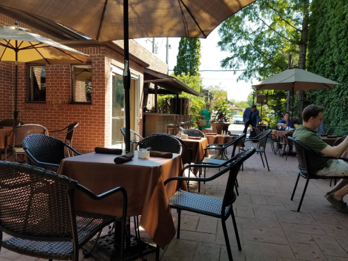 Outdoor café scene with tables, umbrellas, and patrons enjoying the ambiance in a green, shaded area.