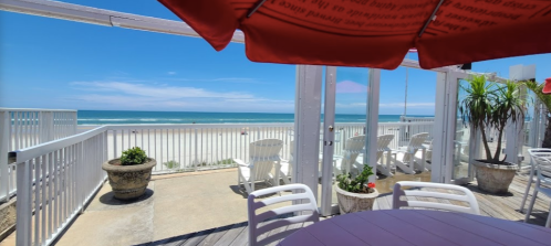 A sunny beach view from a patio with white chairs and a red umbrella, overlooking the ocean and sandy shore.