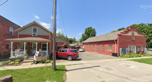 A red brick building and a house with a porch, set on a sunny street with parked cars and greenery.