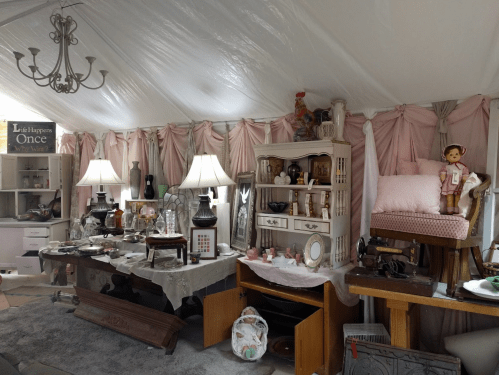 A cozy indoor space filled with vintage furniture, decor, and collectibles, featuring lamps, a doll, and pink drapery.