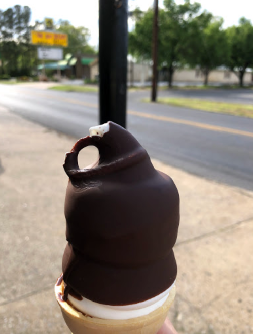 A hand holding a chocolate-dipped ice cream cone with a blurred street and trees in the background.