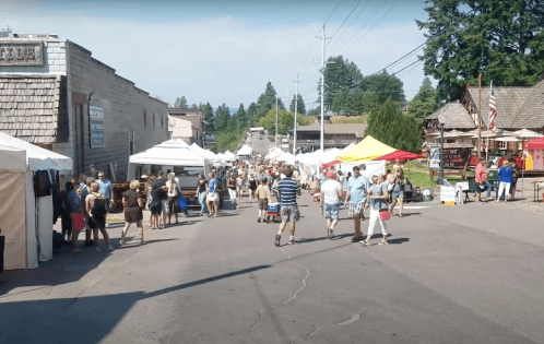 A bustling street market with tents, vendors, and crowds of people enjoying the sunny day. Trees and buildings in the background.