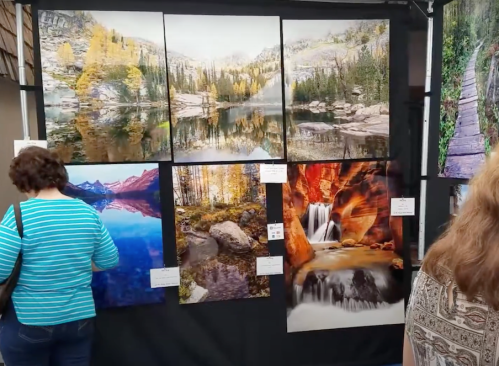 A gallery display featuring six landscape photographs of mountains, lakes, and waterfalls, with visitors admiring the art.