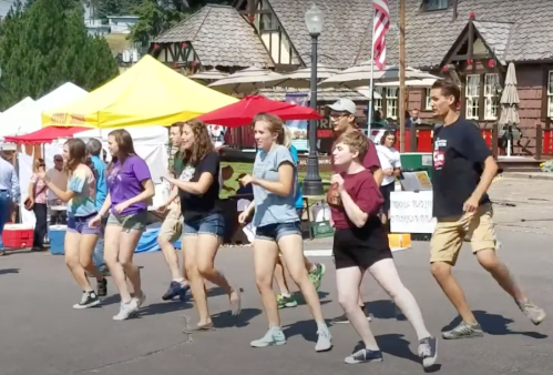 A group of young people dancing in a line at an outdoor festival, with colorful tents in the background.