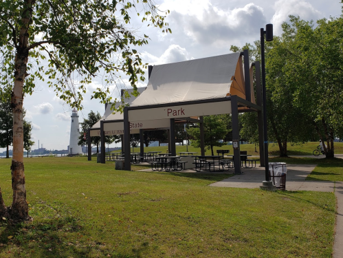 A park pavilion with picnic tables, surrounded by grass and trees, with a lighthouse visible in the background.