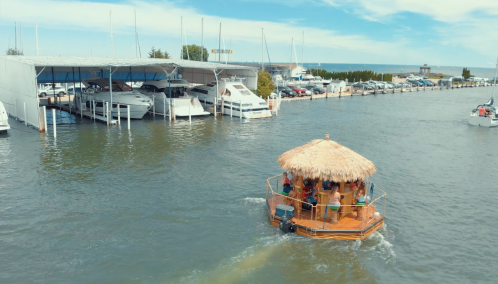 A tiki-style boat with people on it navigates a marina filled with larger boats and a clear blue sky.