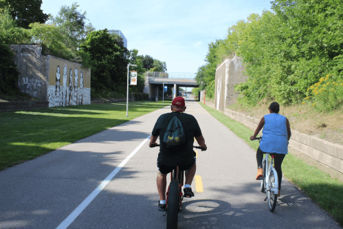 Two cyclists ride on a paved path surrounded by greenery and murals, with a bridge in the background.