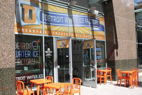 Exterior of the Detroit Water Ice Factory, featuring bright orange tables and a sign for breakfast treats and sweets.