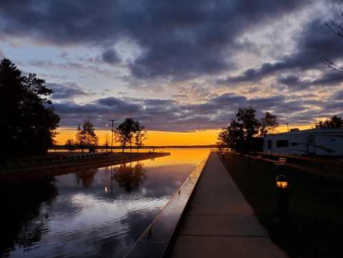 A serene sunset over a calm waterway, with trees and a pathway lined by soft clouds and warm orange hues.