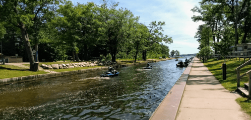 A serene canal scene with boats cruising along the water, lined by trees and a pathway on the right.