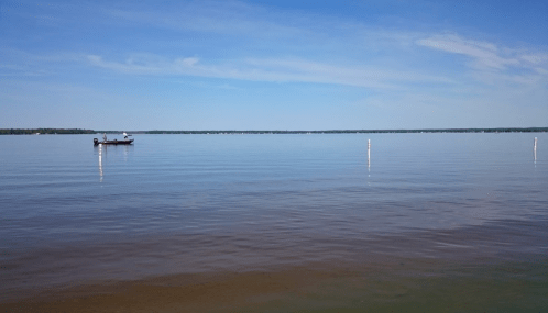 A calm lake under a clear blue sky, with a small boat in the distance and white markers in the water.