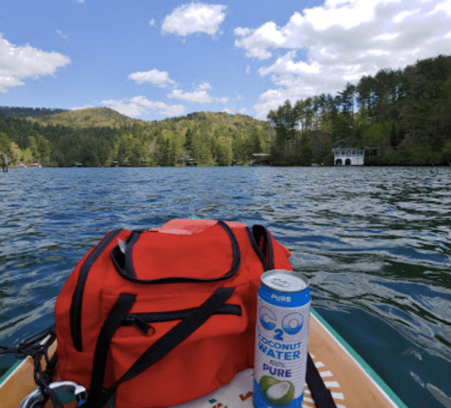 A red backpack and a can of coconut water on a paddleboard, with a scenic lake and forested hills in the background.