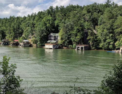 A serene lake scene with houses along the shore, surrounded by lush green trees under a partly cloudy sky.