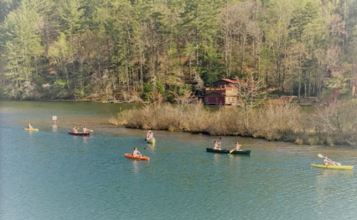 A serene lake scene with several kayakers paddling near a wooded shore and a red cabin in the background.