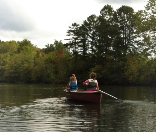 Two people rowing a small boat on a calm river surrounded by trees in autumn colors.