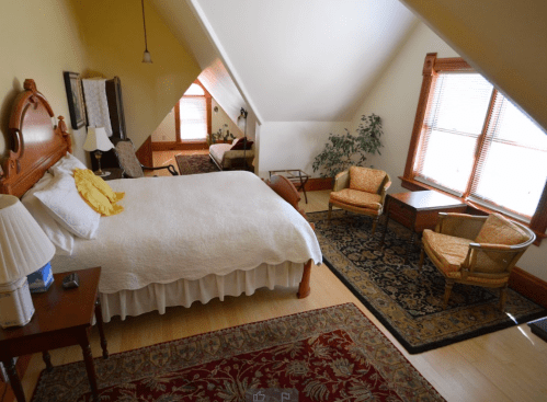 Cozy attic bedroom with a bed, two chairs, a table, and large windows, featuring warm colors and decorative rugs.