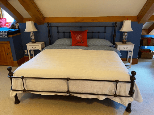 A cozy bedroom featuring a metal bed frame, blue walls, and two bedside tables with lamps. A decorative red pillow is on the bed.