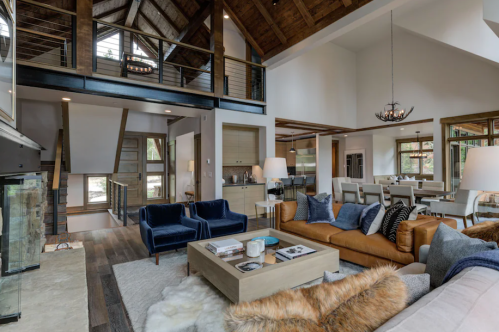 A spacious, modern living room with high ceilings, wooden beams, and stylish furniture, featuring a cozy fireplace.