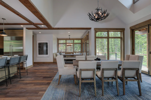 Modern dining area with a wooden table, chairs, and large windows overlooking a forested view. Cozy and inviting atmosphere.