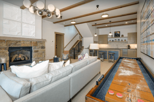 Cozy living area with a fireplace, modern furniture, and a shuffleboard table, featuring a stylish kitchen in the background.
