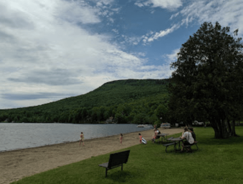 A sandy beach by a lake with people enjoying the water, surrounded by green hills and a cloudy sky.