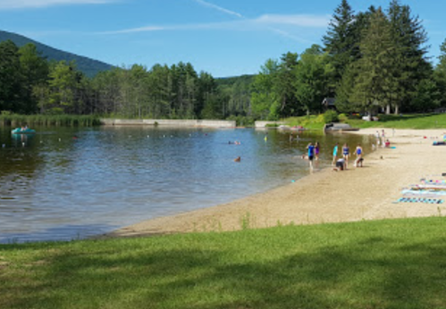 A sandy beach by a calm lake, with people walking and enjoying the outdoors surrounded by trees and mountains.
