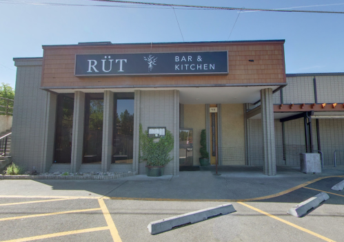 Exterior of Rüt Bar & Kitchen, featuring a modern facade with large windows and a wooden sign. Clear blue sky above.