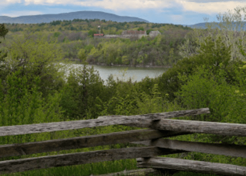 A scenic view of a river surrounded by lush greenery and distant mountains under a cloudy sky. A wooden fence in the foreground.