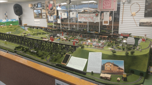 A detailed model train display featuring a miniature town, landscape, and bridges in a museum setting.
