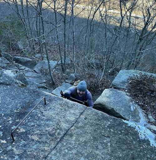 A climber ascends a rocky surface, surrounded by bare trees and a distant view of a valley.