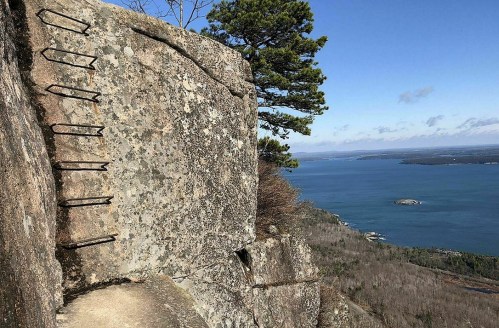 A rocky cliff with metal rungs leading up, overlooking a scenic view of a lake and distant islands.