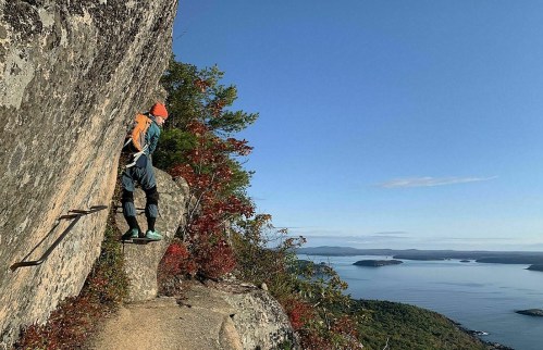 A climber ascends a rocky cliff with a scenic view of water and trees in the background on a clear day.