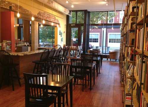 Cozy café interior with wooden tables, chairs, a bar, and bookshelves, featuring large windows and warm lighting.