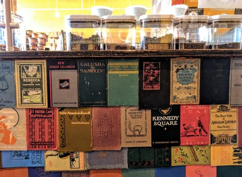 A colorful display of vintage books on a shelf, with jars and a café setting in the background.