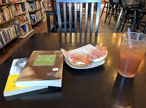 A table in a café with a book, a sandwich with ham, and a drink, surrounded by bookshelves in the background.