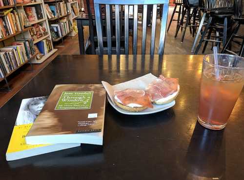 A table in a café with a book, a sandwich with ham, and a drink, surrounded by bookshelves in the background.