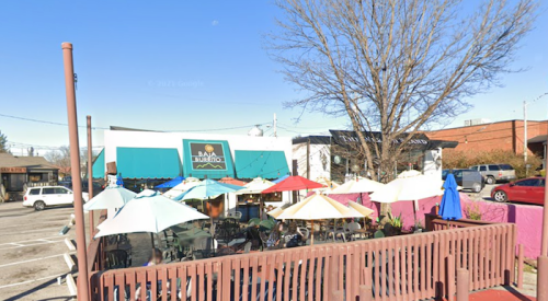 Outdoor dining area with colorful umbrellas, surrounded by trees and buildings on a clear day.