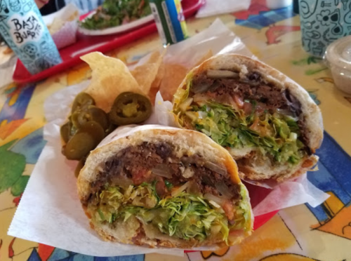 A close-up of a sandwich filled with lettuce, tomatoes, and meat, served with chips and jalapeños on the side.