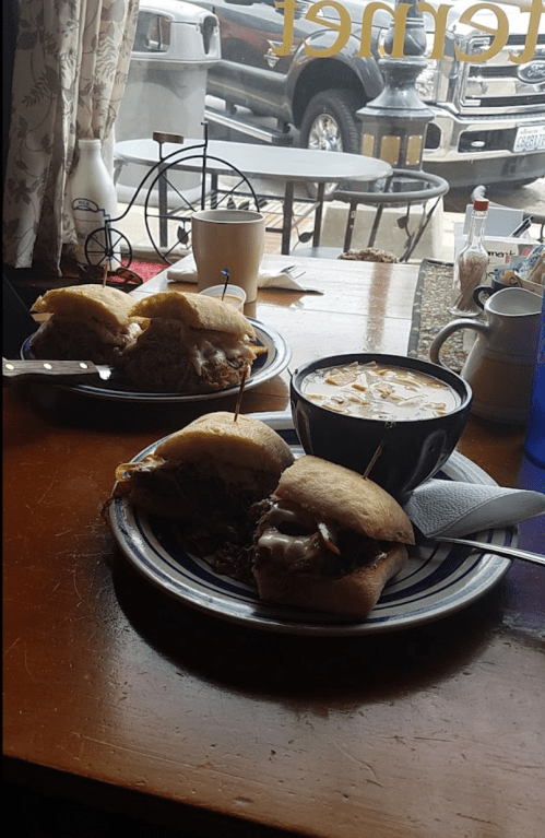 Two sandwiches on a plate with a bowl of soup, set on a wooden table with a view of a street outside.
