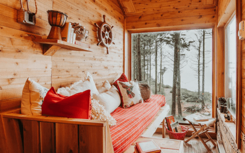 Cozy wooden cabin interior with a red sofa, decorative pillows, and a view of trees outside. Warm, inviting atmosphere.