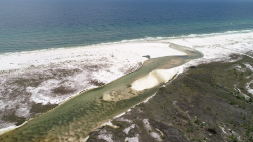 Aerial view of a sandy beach with a winding river flowing into the ocean, surrounded by greenery and clear blue water.