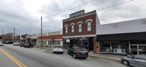 A street view of a small town with shops, parked cars, and a cloudy sky.