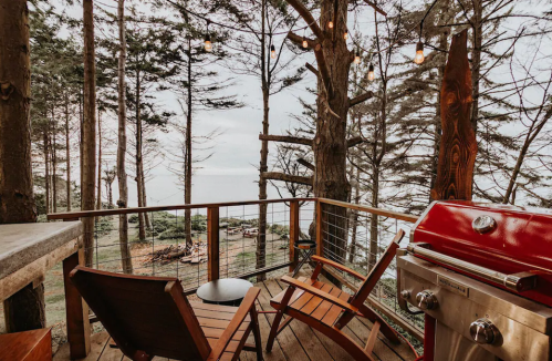 A cozy deck with wooden chairs, a small table, and a red grill, surrounded by trees and overlooking a serene ocean view.
