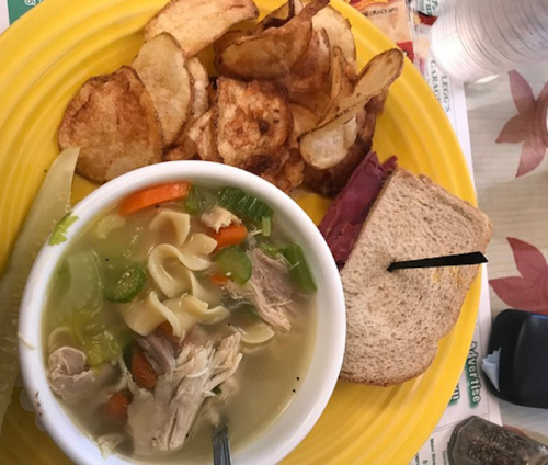 A plate with chicken noodle soup, potato chips, and a sandwich on whole grain bread with a pickle on the side.